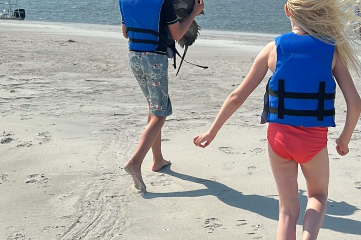Two children in blue life vests walk on a sandy beach near water.