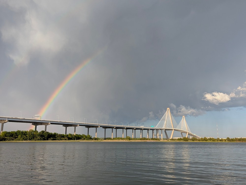 Bridge over water with a rainbow in cloudy sky.