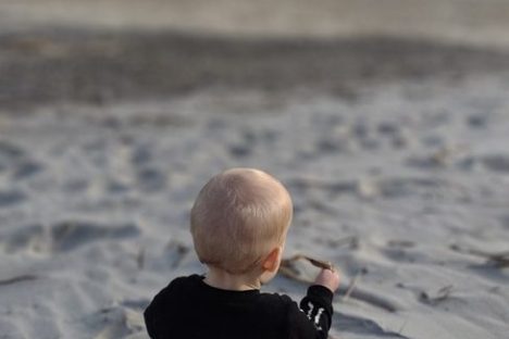 Child sitting on sandy beach, facing the ocean, wearing black and holding a stick.