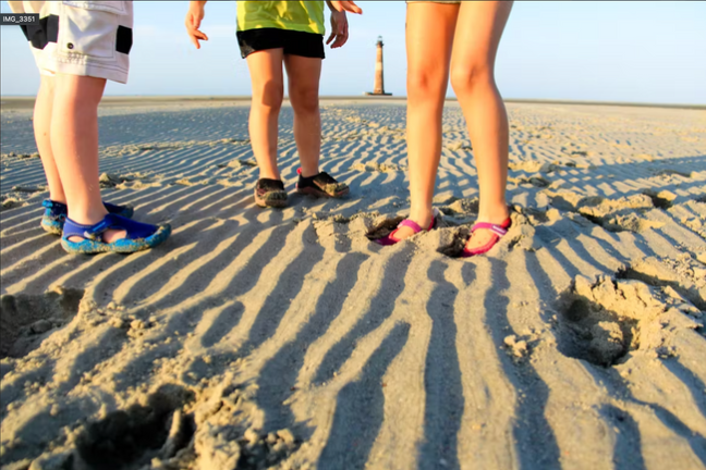 Three children in colorful shoes on a rippled sand beach with a distant lighthouse.
