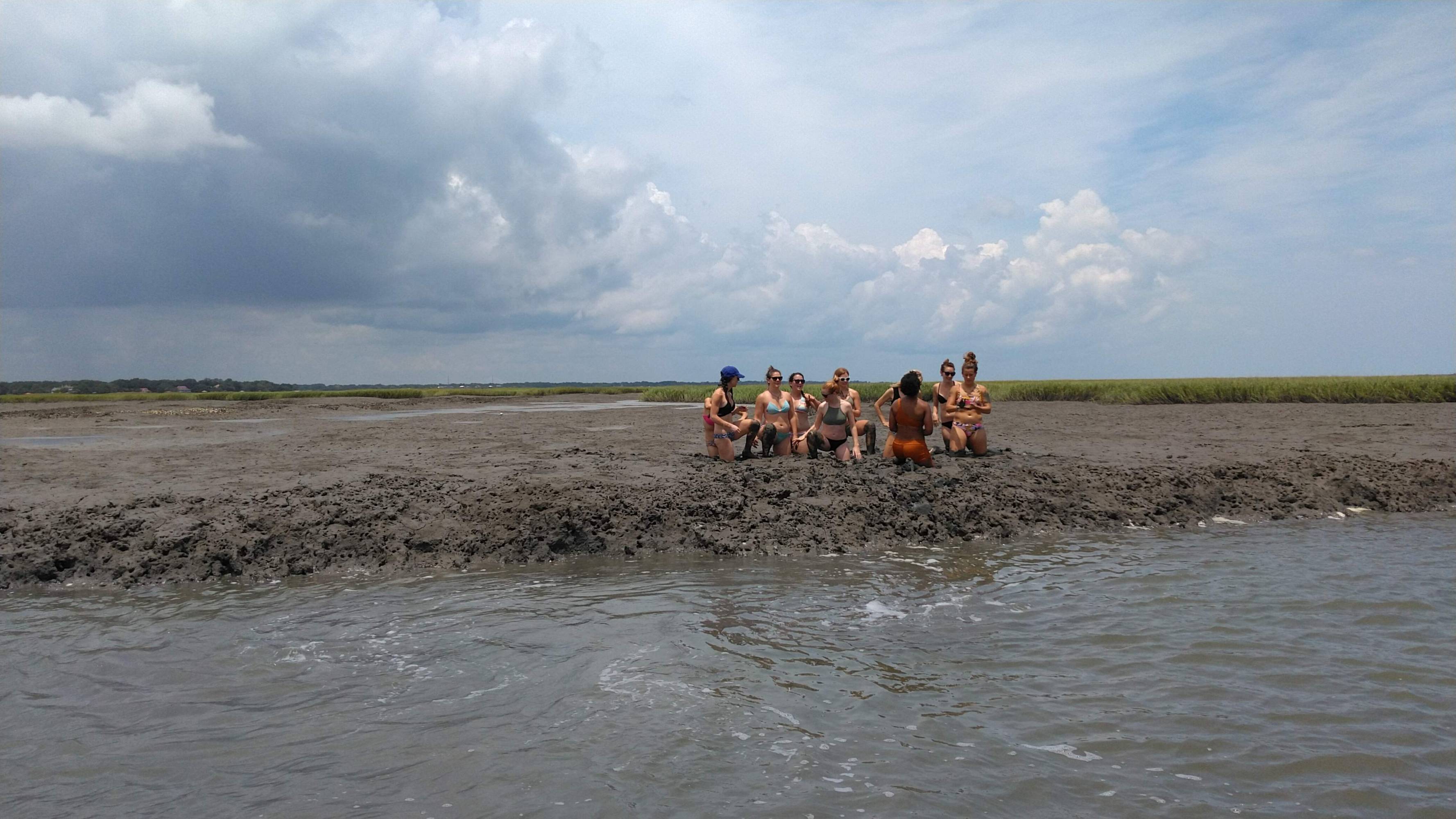 Group of people sitting on muddy ground by the water under a cloudy sky.