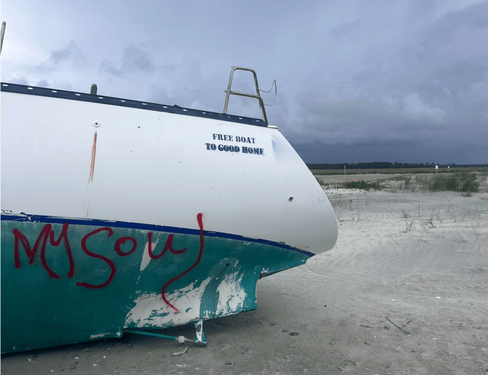 Abandoned boat on sandy beach with graffiti and 'Free boat to good home' text.