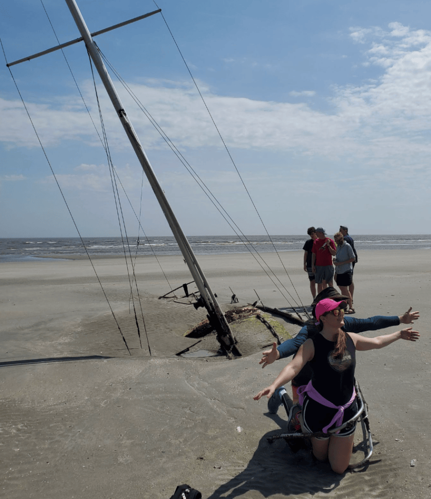 People pose on a sandy beach near a partially buried sailing mast.